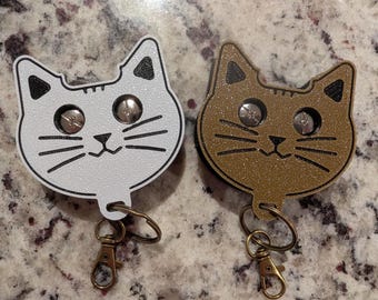 May include: Two cat-shaped keychains, one white and one bronze, each featuring a textured surface, black engraved facial details, two silver-toned screw eyes for eyes, and a bronze-toned metal lobster clasp attached to a small ring at the base.