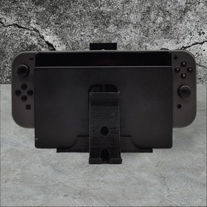 Puede incluir: Un soporte de consola Nintendo Switch negro con una base de plástico negro y un soporte de plástico negro. La consola está en posición vertical y el soporte la sostiene.