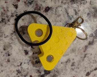 May include: A bright yellow, triangular, textured plastic keychain shaped like a slice of Swiss cheese with circular indentations. It features two embedded silver-colored circular magnets and a black elastic hair tie, attached to a brass-toned metal lobster clasp and keyring.