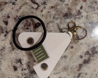 May include: A white triangular plastic hair tie holder with a textured surface, featuring two circular silver magnets and a central green rectangular insert. A black elastic hair tie is looped around the holder, which includes a gold-toned metal lobster clasp keychain attachment.