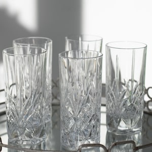 May include: Five clear, tall drinking glasses with a cut glass design, arranged on a silver-toned metal tray. The glasses have a vertical, textured pattern. The tray has a reflective surface.