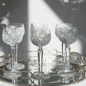 May include: Five clear glass goblets with intricate cut designs sit on a silver tray with a mirrored surface. The goblets are arranged in a circle, with the stems pointing towards the centre of the tray.