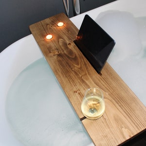 Puede incluir: Una bandeja de baño de madera con un soporte integrado para teléfono o tableta, un vaso para bebidas y dos velas. La bandeja está colocada en el borde de una bañera blanca llena de agua y burbujas.