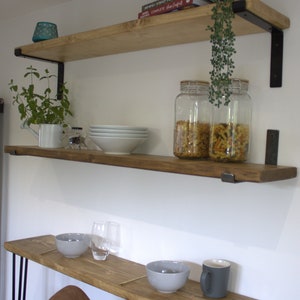 May include: Two wooden shelves with black metal brackets, mounted on a white wall. The top shelf has a cookbook, a small potted plant, and two glass jars filled with pasta. The bottom shelf has a white bowl, a small watering can, and a small container of spices.