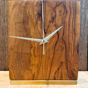 Puede incluir: Un reloj de madera cuadrado con un acabado de vetas de madera natural. La esfera del reloj presenta manecillas plateadas y un diseño minimalista. La base es de metal dorado. Un acento decorativo para un escritorio o estante.