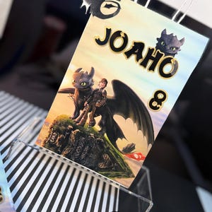 May include: A white paper gift bag featuring a colorful illustration of characters from the "How to Train Your Dragon" movie. The bag has the name "JOAHO" and the number "8" printed on it. The bag has white paper handles and a black dragon cutout.