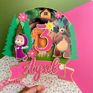 May include: A circular cake topper featuring a forest scene with a wooden house, a cartoon girl in a purple dress, and a brown bear. The number 3 is displayed in pink with a yellow sun icon. A pink banner at the bottom displays the name Aysel in white.