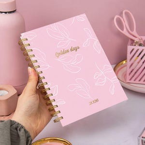 Puede incluir: Una agenda rosa con espiral y detalles dorados, con el texto "Golden days" y "2026" en dorado. La portada presenta ilustraciones de hojas blancas. La agenda está sujeta con una mano.