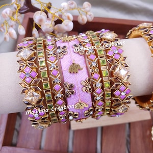 Könnte beinhalten: Eine Sammlung verzierter Armreifen in Gold-, Rosa- und Grüntönen. Die Armreifen weisen geometrische und florale Designs mit funkelnden Akzenten auf. Der zentrale Armreif ist helllila mit einem Elefantenanhänger und dem Text "Mayilina Creations".