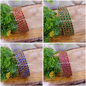 Op de afbeelding: Vier gouden armbanden met vierkante rode, groene, blauwe en roze edelstenen. De armbanden zijn op elkaar gestapeld.