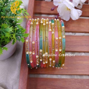 May include: A set of vibrant bangles in a rainbow of colours, including pink, green, yellow, and blue. The bangles are decorated with gold-tone details and small, pearl-like beads. The text "Mayilina Creations" is displayed.