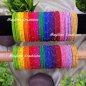 Puede incluir: Una pila de pulseras de hilo de seda de colores con cuentas doradas. Las pulseras están dispuestas en forma de arcoíris, cada pulsera es de un color diferente. El texto "Mayilna Creations" es visible en la imagen.