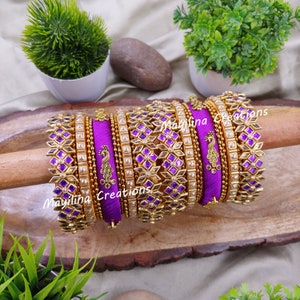May include: A set of purple and gold bangles with intricate designs. The bangles feature a geometric pattern with purple accents and a peacock design. The text "Mayilina Creations" is visible.