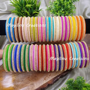 以下が含まれることがあります： 青、緑、ピンク、黄色など、さまざまな色合いのカラフルなバングルのコレクション。各バングルには、小さな輝く装飾が並んでいます。「Mayilina Creations」の文字がバングルに表示されています。