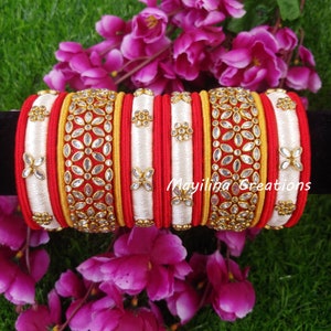 May include: A set of red and white silk thread bangles with gold accents and clear rhinestone embellishments. The bangles are stacked on top of each other and feature a floral design. The text "Mayiliha Creations" is visible on the bottom right of the image.