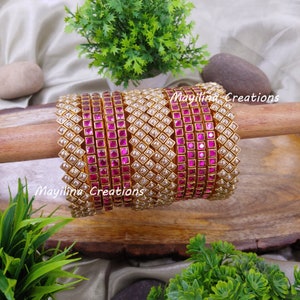 Könnte beinhalten: Ein Set aus 10 goldenen Armreifen mit rosa und weißen Steinen. Die Armreifen sind auf einer Holzoberfläche gestapelt. Mayilina Creations ist auf der Oberfläche geschrieben.