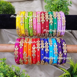 Könnte beinhalten: Ein Stapel bunter Seidenfaden-Armreifen mit goldenen und weißen Verzierungen. Die Armreifen sind in Regenbogenfarben angeordnet, wobei jeder Armreifen ein einzigartiges Design aufweist.