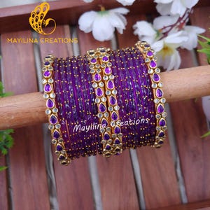May include: A stack of purple bangles with gold accents and teardrop-shaped purple gems. The bangles are displayed on a wooden dowel, with the text "Mayilina Creations" visible. White flowers and green leaves are in the background.