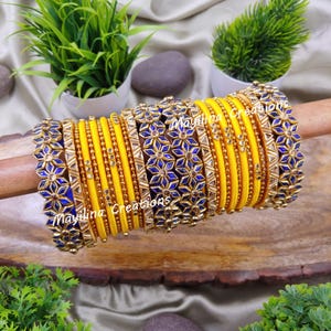Könnte beinhalten: Ein Set aus 16 gelben und blauen Armreifen mit goldenen Akzenten und floralen Designs. Die Armreifen sind auf einer Holzoberfläche gestapelt.