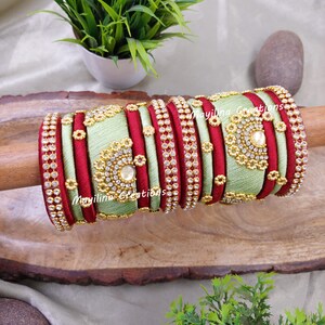 May include: A set of decorative bangles in shades of light green and red, adorned with sparkling rhinestones and gold-colored embellishments. The bangles are arranged on a wooden dowel, showcasing their intricate design and craftsmanship. The text "Mayilina Creations" is visible.