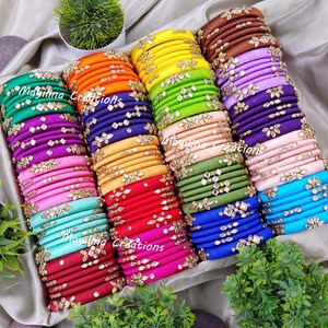 May include: A collection of colorful silk thread bangles with jeweled embellishments. The bangles are stacked in a box and feature a variety of colors, including red, pink, blue, green, and yellow.