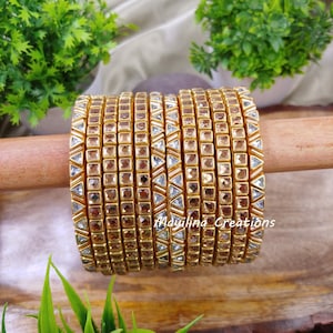 Peut inclure: Une pile de bracelets dorés avec des accents de pierres claires carrées et triangulaires. Les bracelets sont présentés sur une cheville en bois, avec des plantes vertes en arrière-plan. Le texte "Maulina Creations" est visible.