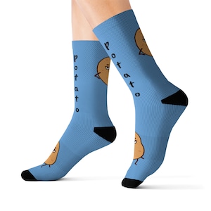 Könnte beinhalten: Hellblaue Socken mit einem Cartoon-Kartoffel-Design. Die Socken zeigen eine braune Kartoffel-Figur mit einem lächelnden Gesicht und dem Wort "potato" vertikal gedruckt. Die Fersen und Zehen sind schwarz.