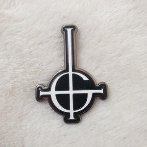 Könnte beinhalten: Eine silberne Metallstifte mit einem schwarzen und weißen umgekehrten Kreuz in einem Kreis. Der Stift ist ein beliebtes Symbol der Band Ghost.