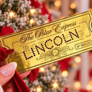 May include: A gold-toned metal ticket ornament featuring the text The Polar Express and ROUND TRIP with a holly and bell graphic. The ticket includes the number 0001225 and is attached to a deep red velvet ribbon for hanging.