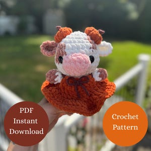 以下が含まれることがあります： 白、茶色、ピンクの配色で手作りのカウのぬいぐるみ。オレンジ色のドレスに茶色のリボンを着ています。画像には「PDF Instant Download」と「Crochet Pattern」という文字も含まれています。