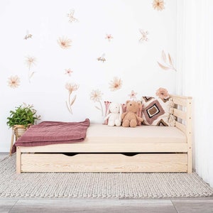 Op de afbeelding: Een licht houten kinderbed met een opberglade. Het bed heeft een beige matras en is versierd met een roze en bruin gepatroneerd kussen en twee knuffels. De muur achter het bed is versierd met bloemenstickers.