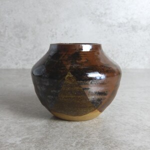 May include: A small, handcrafted ceramic vase with a rounded body and a narrow opening. The vase features a glossy, dark brown glaze with areas of lighter brown and tan, creating a textured appearance. The vase is approximately 10 cm tall.