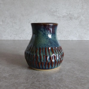 May include: A small, handcrafted ceramic vase with a textured, ribbed design. The vase features a gradient of colours, including deep blue, teal, and brown. The rim is a darker shade of brown. The vase is a decorative item.