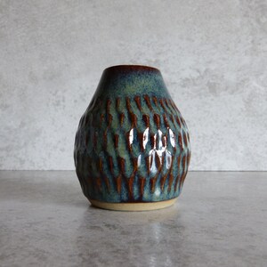 May include: A small, handcrafted ceramic vase with a textured surface. The vase features a teal and brown colour scheme, with a dark brown rim and base. The vase is approximately 13 cm tall and 10 cm wide, with a rounded shape.