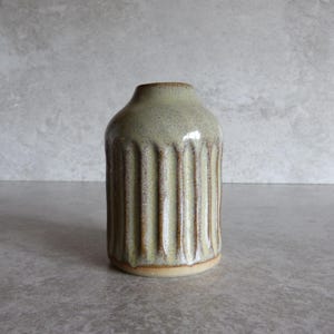 May include: A small, ceramic vase with a light green glaze and vertical ribbed detailing. The vase has a rounded body and a narrow neck. The base is a light tan colour. The vase is approximately 15 cm tall.