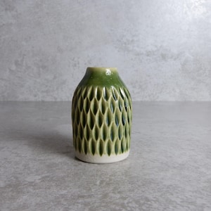 May include: A small, green ceramic vase with a textured, leaf-like pattern. The vase has a white base and a slightly flared top. The vase is approximately 10 cm tall and is suitable for small floral arrangements.