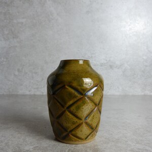 May include: A small, olive-green ceramic vase with a diamond pattern. The vase has a rounded body and a narrow neck. The glaze has a glossy finish. The vase is on a neutral surface.