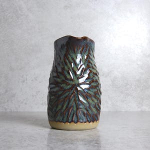 May include: A handcrafted ceramic vase with a textured leaf design. The vase has a dark teal and brown glaze over a beige base. The opening has an irregular shape, enhancing its unique character.