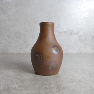 May include: A brown ceramic vase with a rounded body and a narrow neck. The vase has three circular, gold-coloured embellishments. The vase is a decorative item, suitable for displaying flowers or as a standalone art piece.