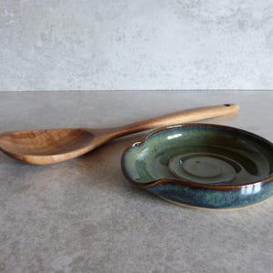 May include: A wooden spoon and a ceramic spoon rest. The spoon is made of light-coloured wood. The spoon rest is oval-shaped with a dark green and blue glaze. The spoon rest is approximately 15 cm long.