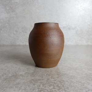 May include: A small, brown ceramic vase with a rounded body and a wide opening. The vase has a textured surface and a simple design. Suitable for displaying flowers or as a decorative accent piece.