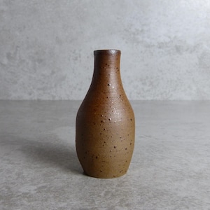 May include: A small, handcrafted ceramic vase with a speckled, brown finish. The vase has a narrow neck and a rounded body, suitable for small flowers or as a decorative accent. The vase is approximately 15 cm tall.