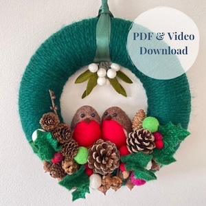 Puede incluir: Una corona de lana verde decorada con petirrojos de fieltro, piñas, acebo y bayas blancas. La corona está colgada en una pared blanca con el texto "PDF & Video Download" en un círculo.