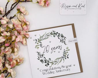 May include: A square wedding anniversary card with a wreath design and the text "20 years". The card also says "Congratulations on your Wedding Anniversary". The card is surrounded by pink and white flowers.
