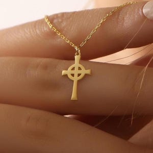 May include: A gold-colored pendant necklace featuring a Celtic cross design. The cross has a distinctive shape with a circular element at the intersection of the arms. The necklace is displayed against a soft, neutral background.