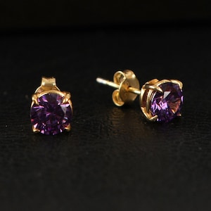 May include: A pair of gold stud earrings with purple gemstones. The earrings have a simple design and are perfect for everyday wear.