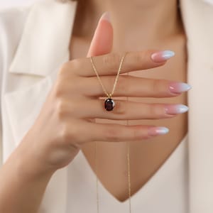 May include: A gold chain necklace with a red oval gemstone pendant. The necklace is being held by a hand with light blue ombre nail polish.