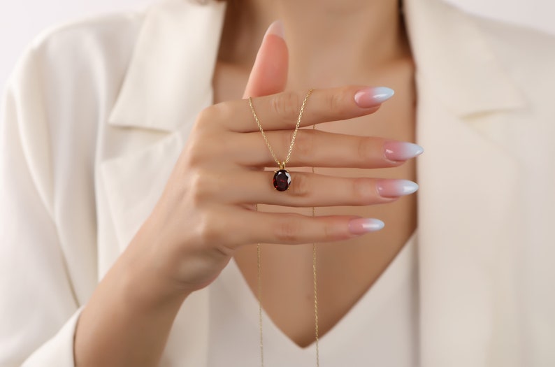 May include: A gold chain necklace with a red oval gemstone pendant. The necklace is being held by a hand with light blue ombre nail polish.