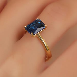 May include: A gold ring featuring a rectangular, deep blue gemstone. The gemstone is set in a four-prong setting, and the band is a polished gold color. The ring is shown on a person's finger.