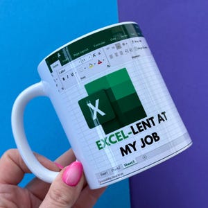 May include: White ceramic mug with an Excel interface design and the text "EXCEL-LENT AT MY JOB". The mug has a white handle. The background is a combination of blue and purple.